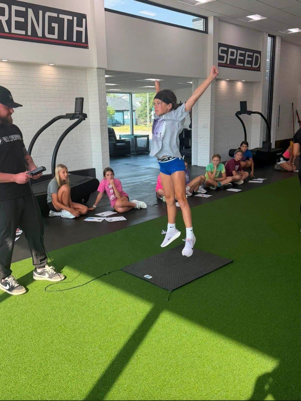 Young athlete jumping off the jump mat while a coach holds the readout and the group watches